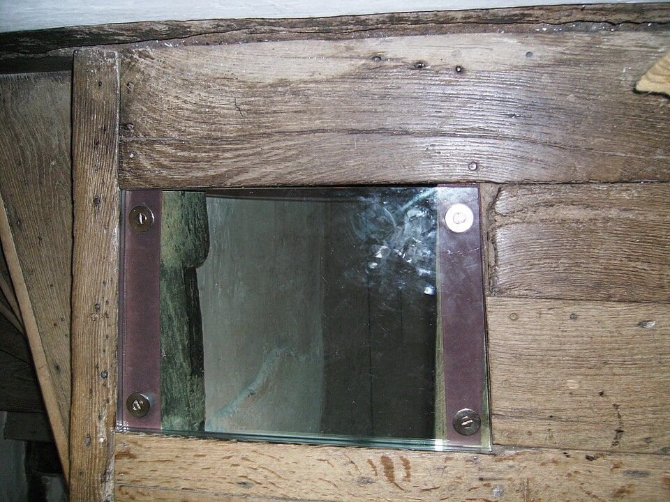 Small rectangular glass inspection window set into rough wooden planks, held by metal strips and screws, showing a dim interior space behind.