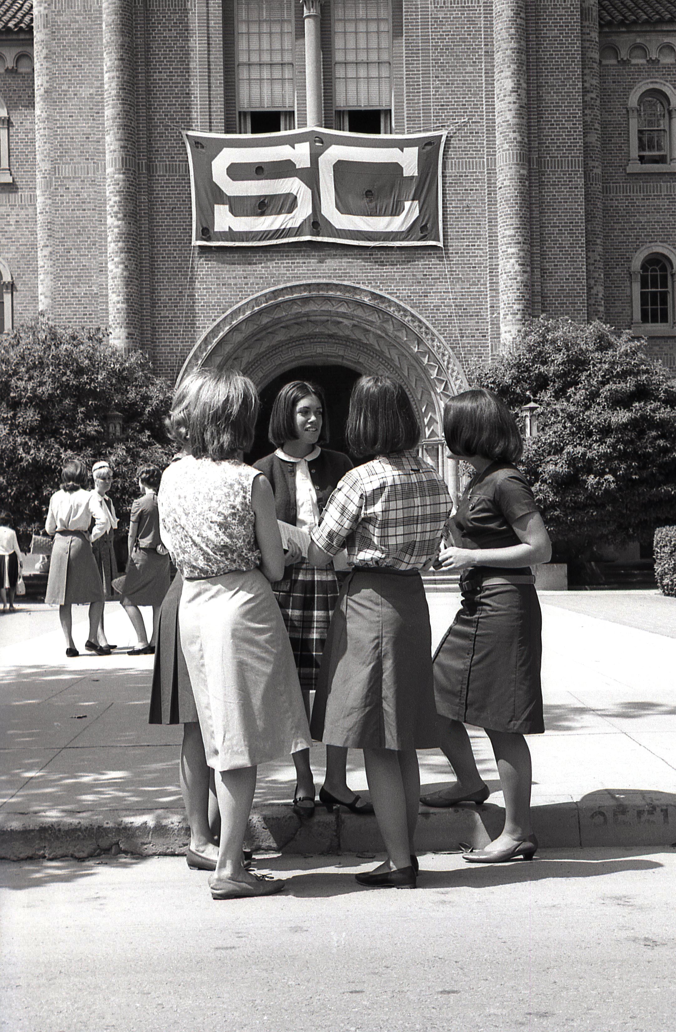 Black-and-white photo of female students talking outside a brick university building with a large “SC” banner above an arched entrance.