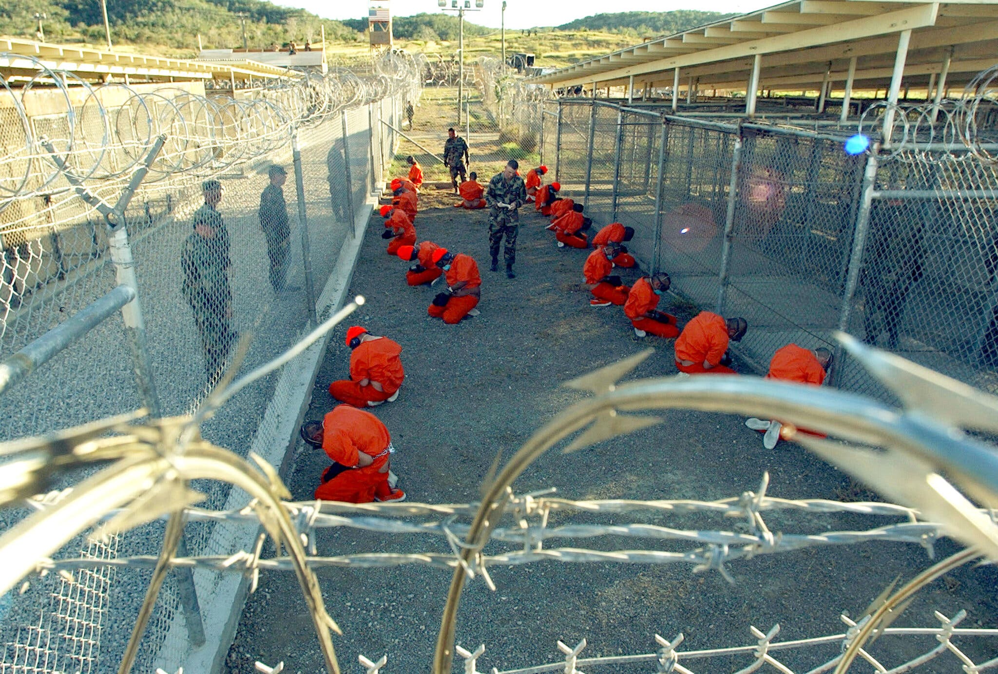 Detainees in orange jumpsuits kneel in a barbed-wire enclosure as armed soldiers stand guard and patrol between fenced cages outdoors