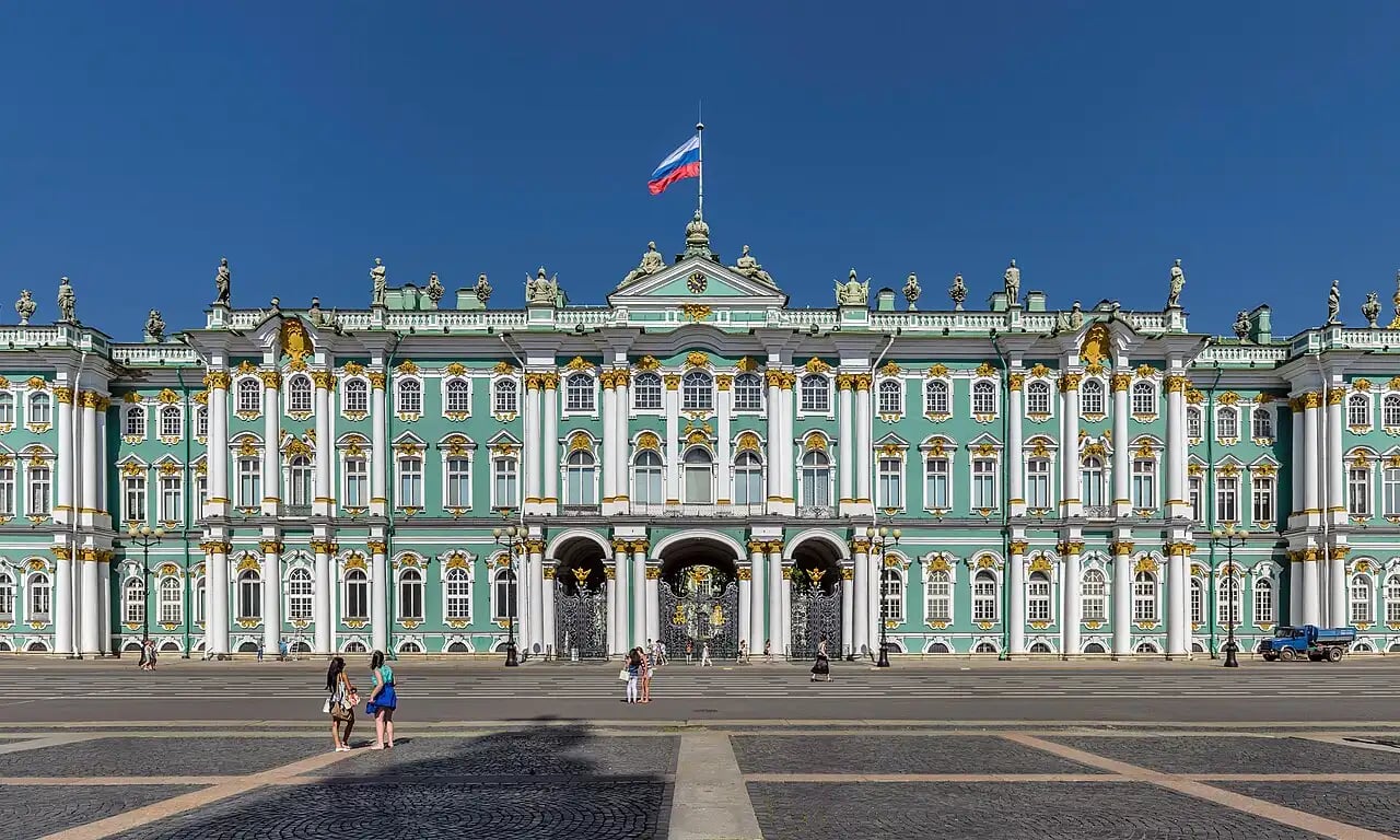 Baroque turquoise palace with white columns and gold trim, Russian flag flying on roof, tourists walking across wide cobbled square under clear blue sky