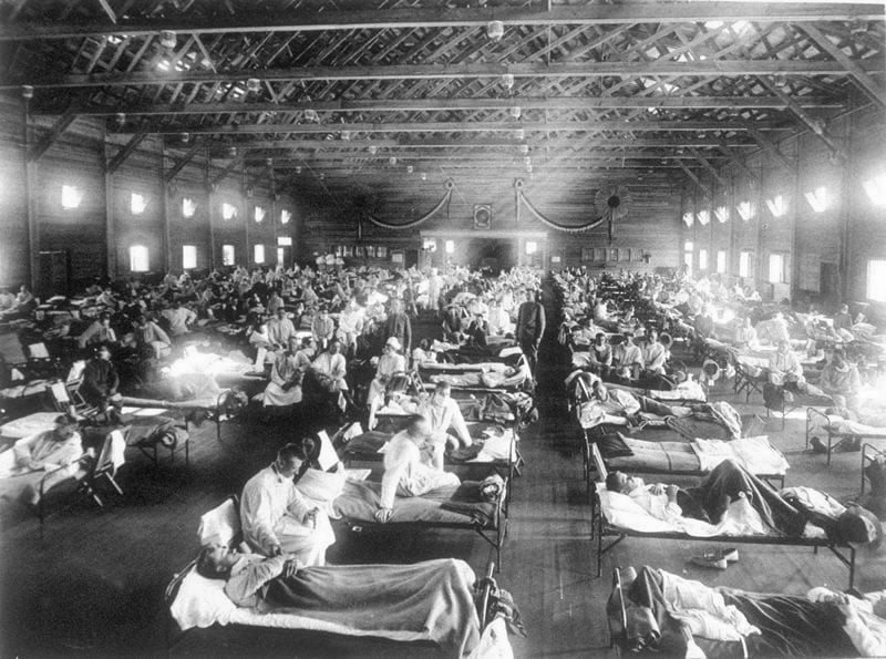 Historic ward filled with rows of camp beds where nurses and doctors tend numerous patients in a large wooden hall during an influenza epidemic