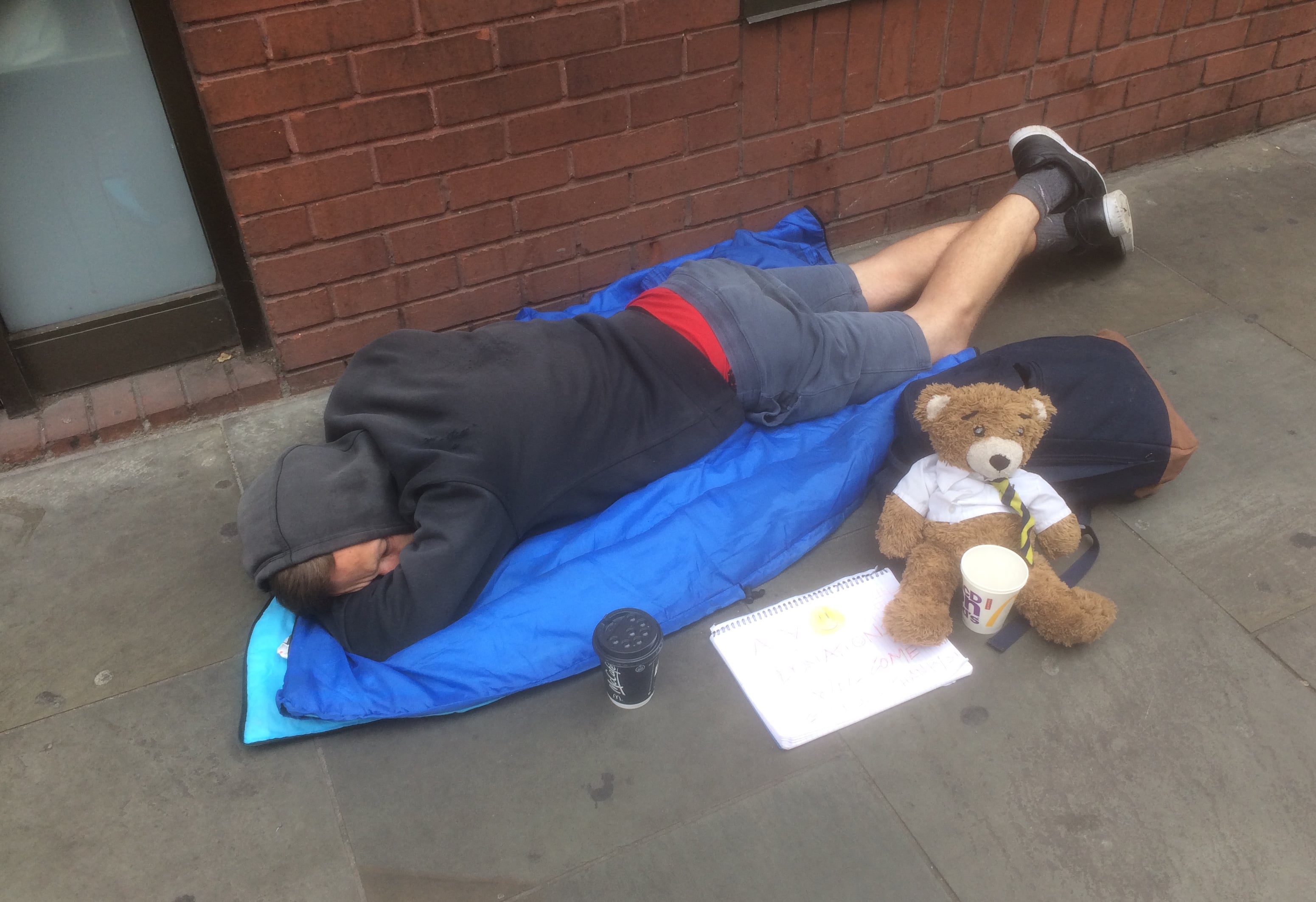 A homeless person sleeps on a sleeping bag on a pavement with a teddy bear and cup for donations