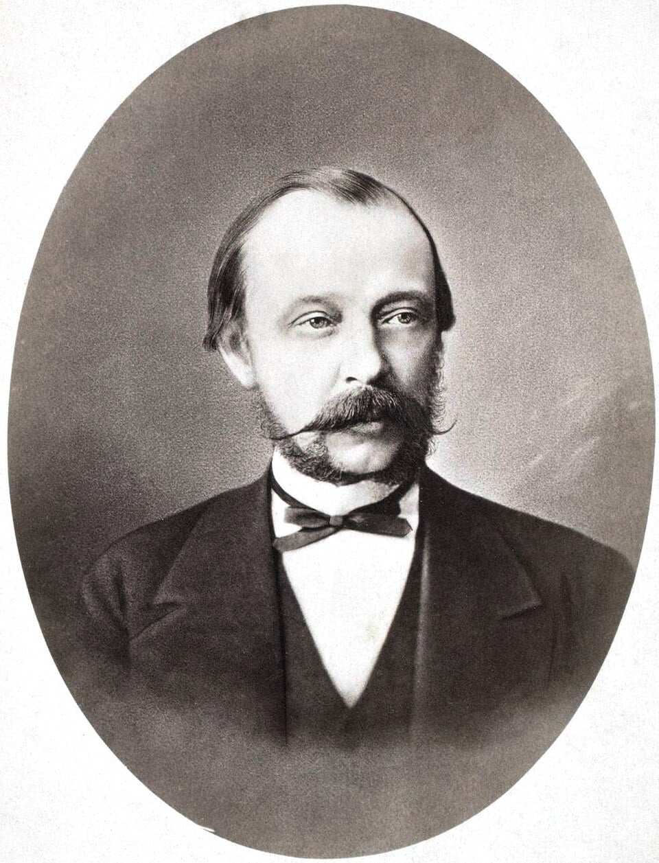 Oval vintage portrait of a middle-aged man with a large moustache and sideburns, wearing a dark suit, waistcoat and bow tie, facing slightly left