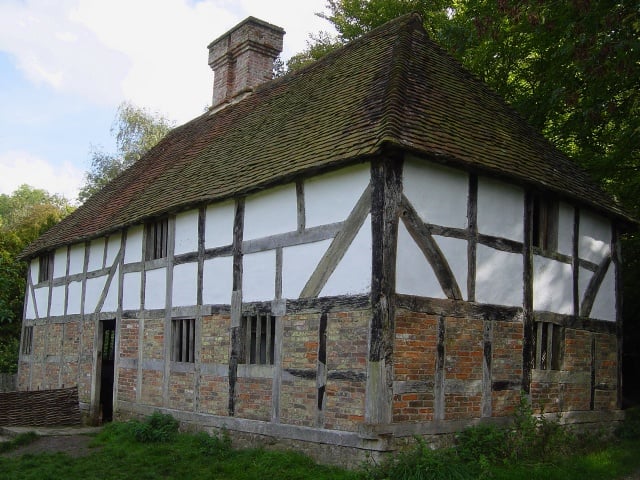 Historic timber-framed cottage with white plaster panels, brick lower walls and a steep tiled roof, set among trees and greenery in the countryside