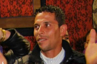 Man with short curly hair and leather jacket smiles and looks sideways in a dim indoor setting with red patterned wall and blurred people around him