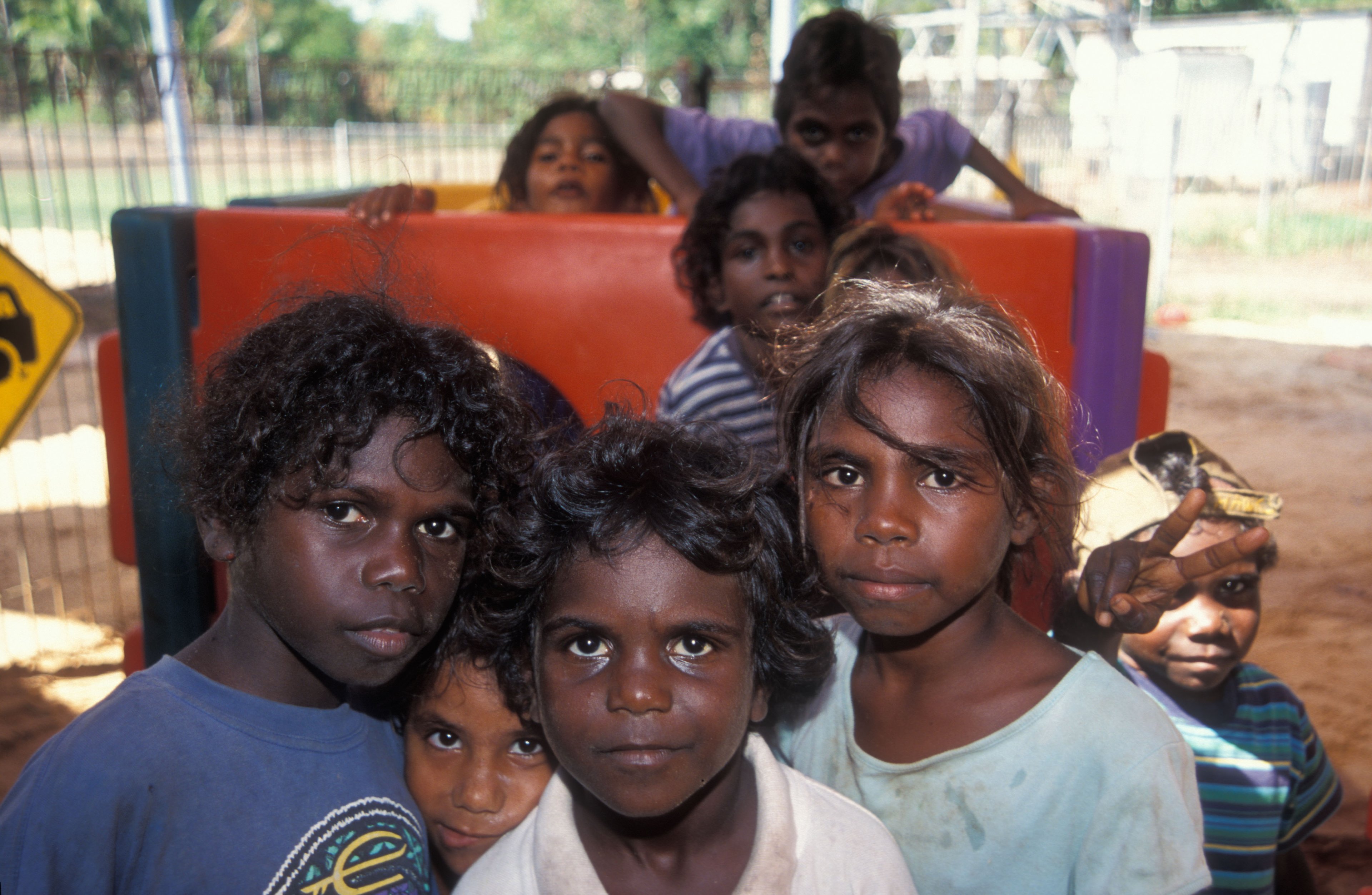 Group of Aboriginal children outdoors, some looking directly at the camera, one making a peace sign, standing in front of colourful playground equipment