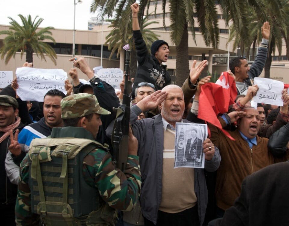 Crowd of protesters with raised arms and signs confront a soldier in camouflage; one man salutes while holding a black‑and‑white portrait.