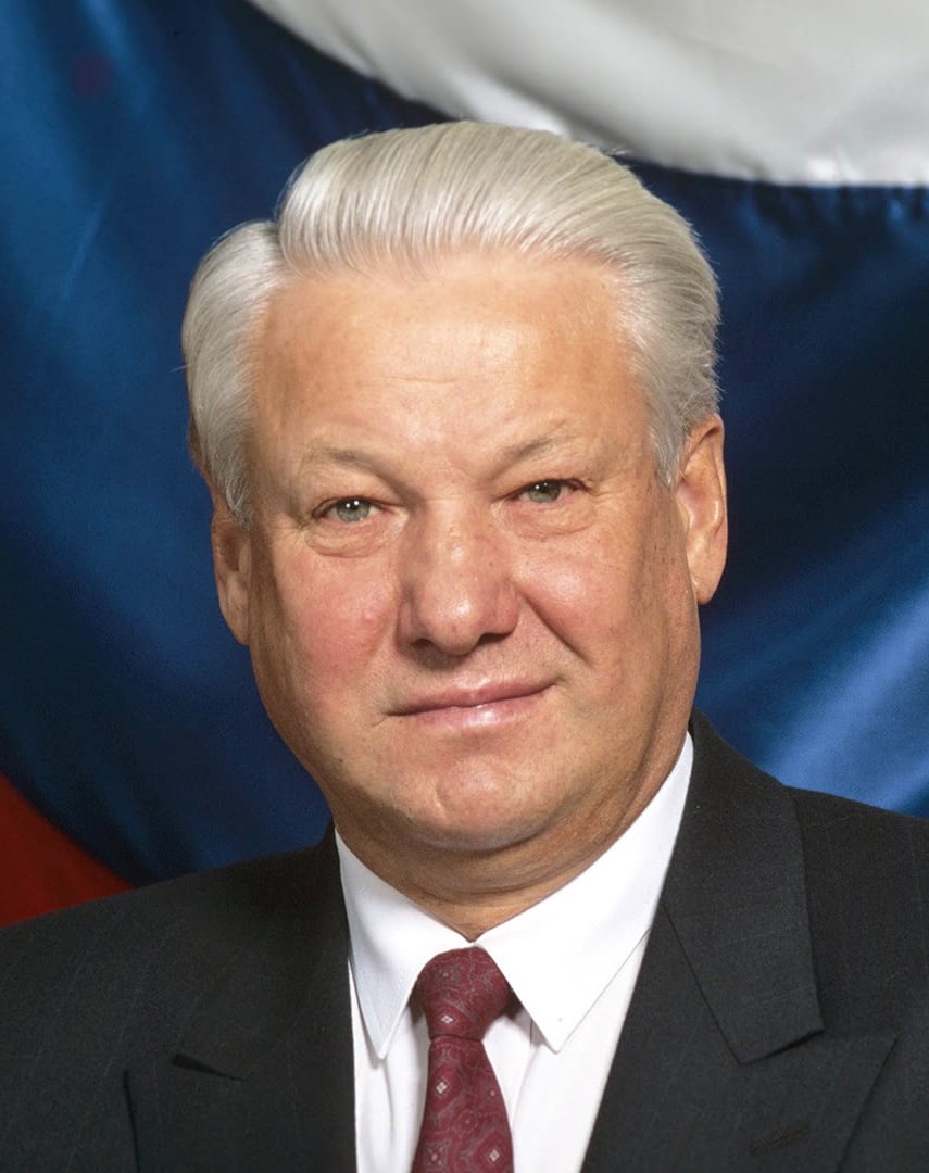 Older man in a dark suit and patterned red tie, with neatly combed white hair, posing formally against a blurred background of red, white and blue fabric