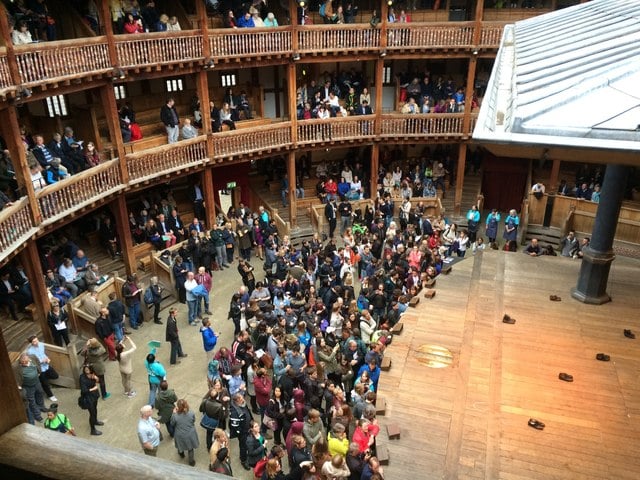 Crowd of visitors standing in the open yard of a wooden, circular Elizabethan-style theatre, with tiered balconies and a large empty stage.