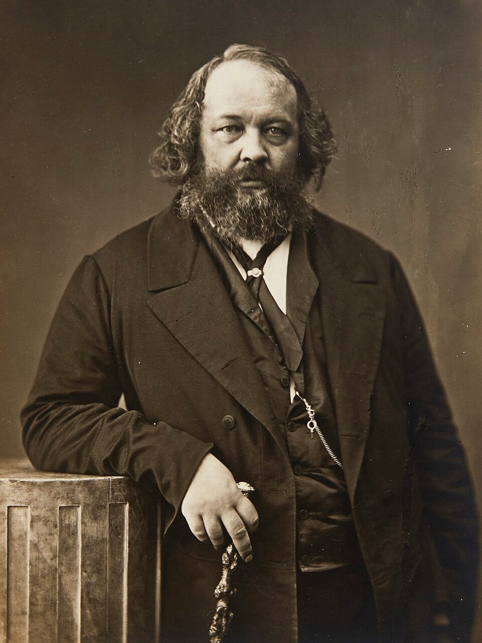 Sepia portrait of a bearded middle-aged man in a dark Victorian suit, standing with one hand on a walking stick and the other resting on a wooden pedestal