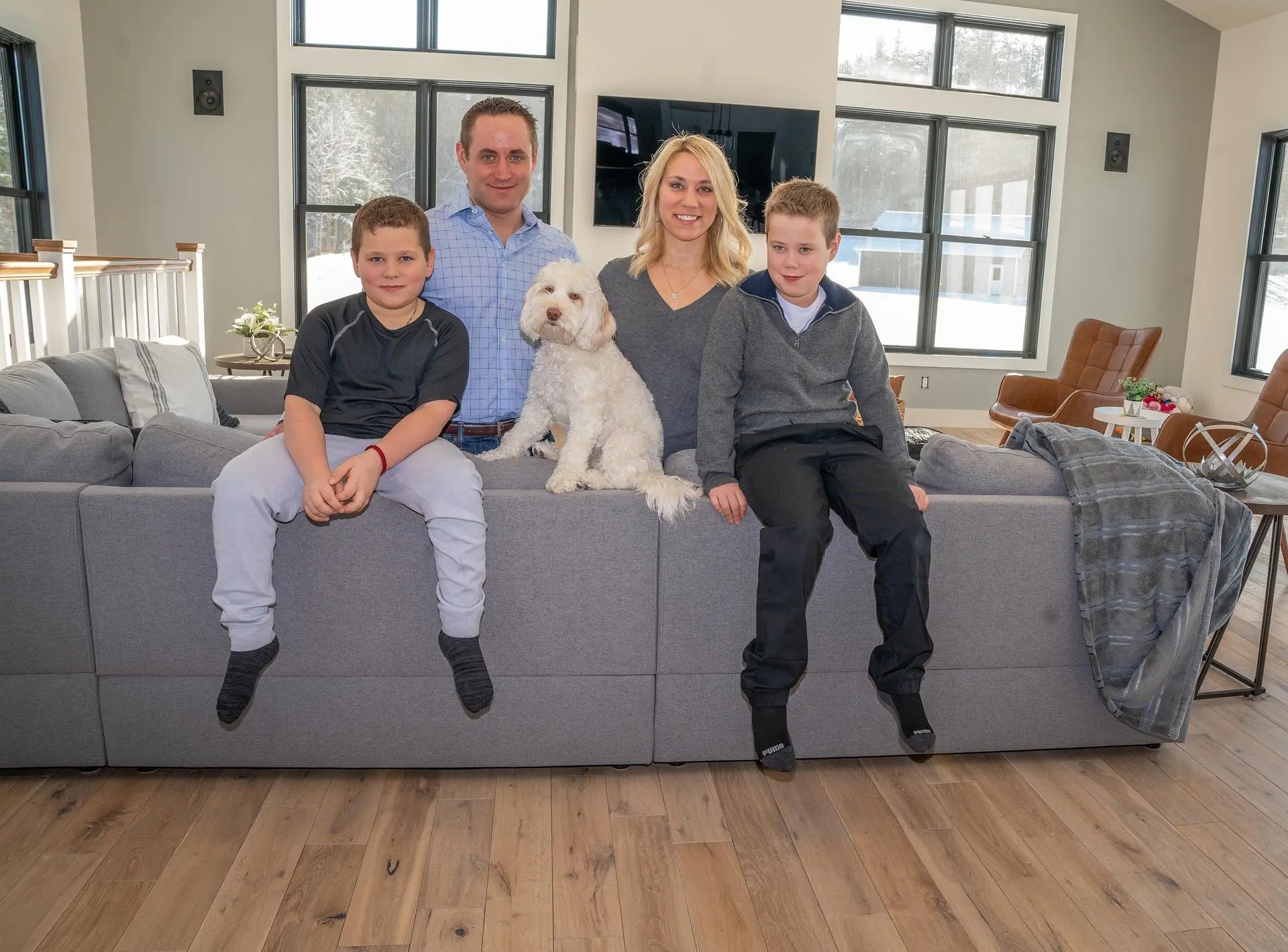 Smiling family with two young boys and a white dog sit on a grey sofa in a bright modern living room with large windows and wooden floor.