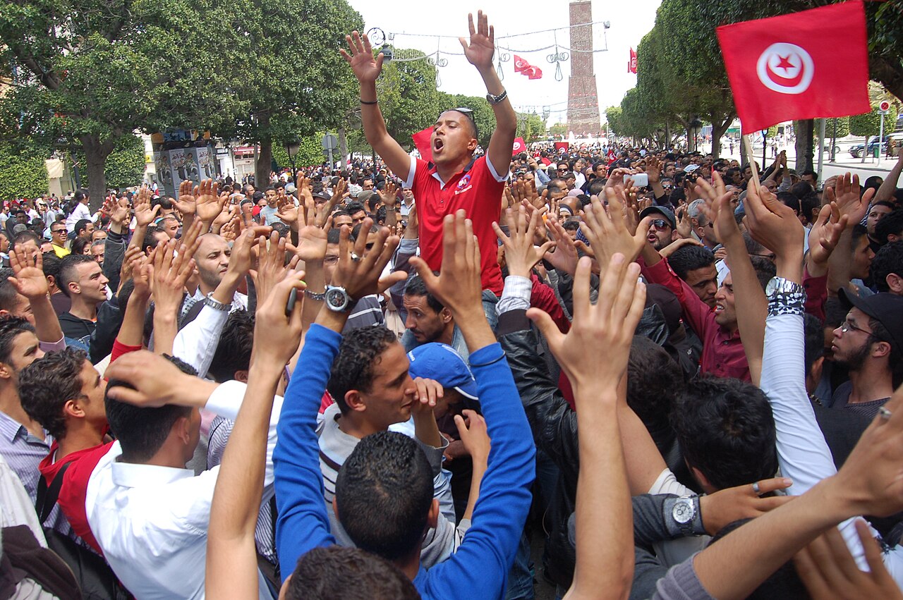 Large crowd of Tunisian protesters with raised hands fills a tree-lined street, a man in red shirt shouting above them and a Tunisian flag waving on the right