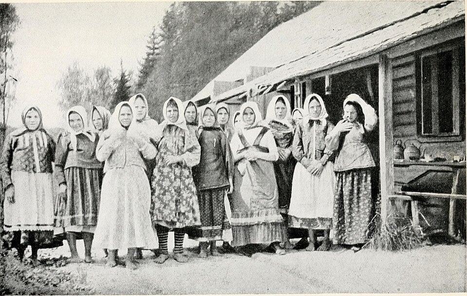 Vintage photo of rural women in headscarves and long skirts standing in a row beside a wooden farmhouse in a forested countryside setting