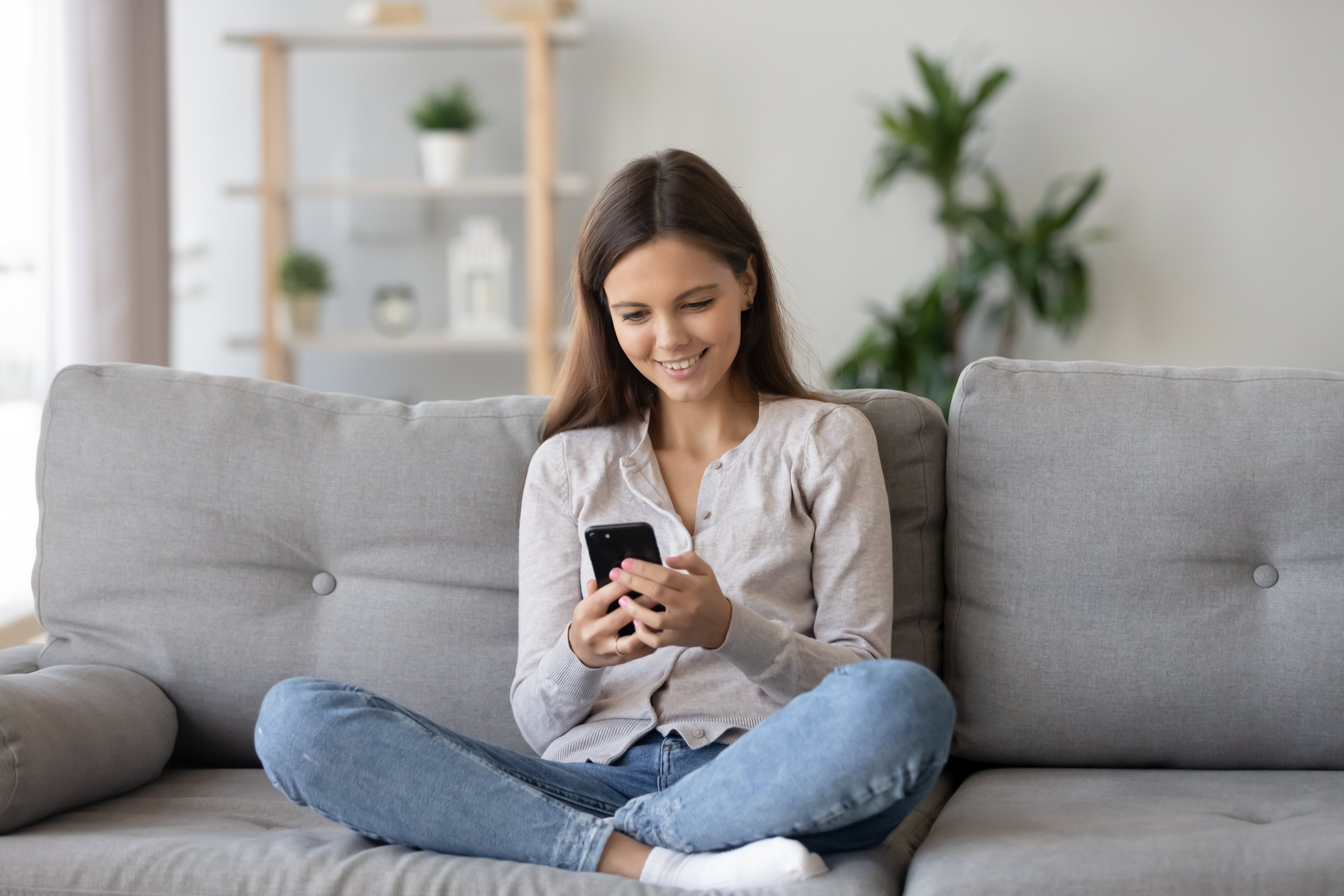 Young woman sitting cross-legged on a grey sofa in a bright living room, smiling while using her smartphone, with shelves and plants in the background