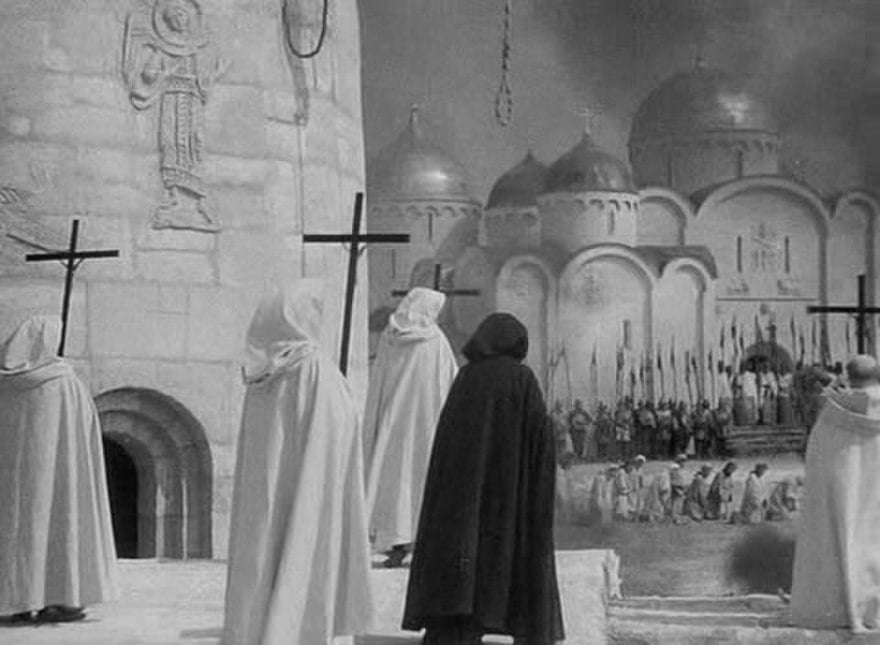 Medieval cloaked figures with tall crosses watch a distant crowd before domed church buildings amid smoke in a dramatic black-and-white scene
