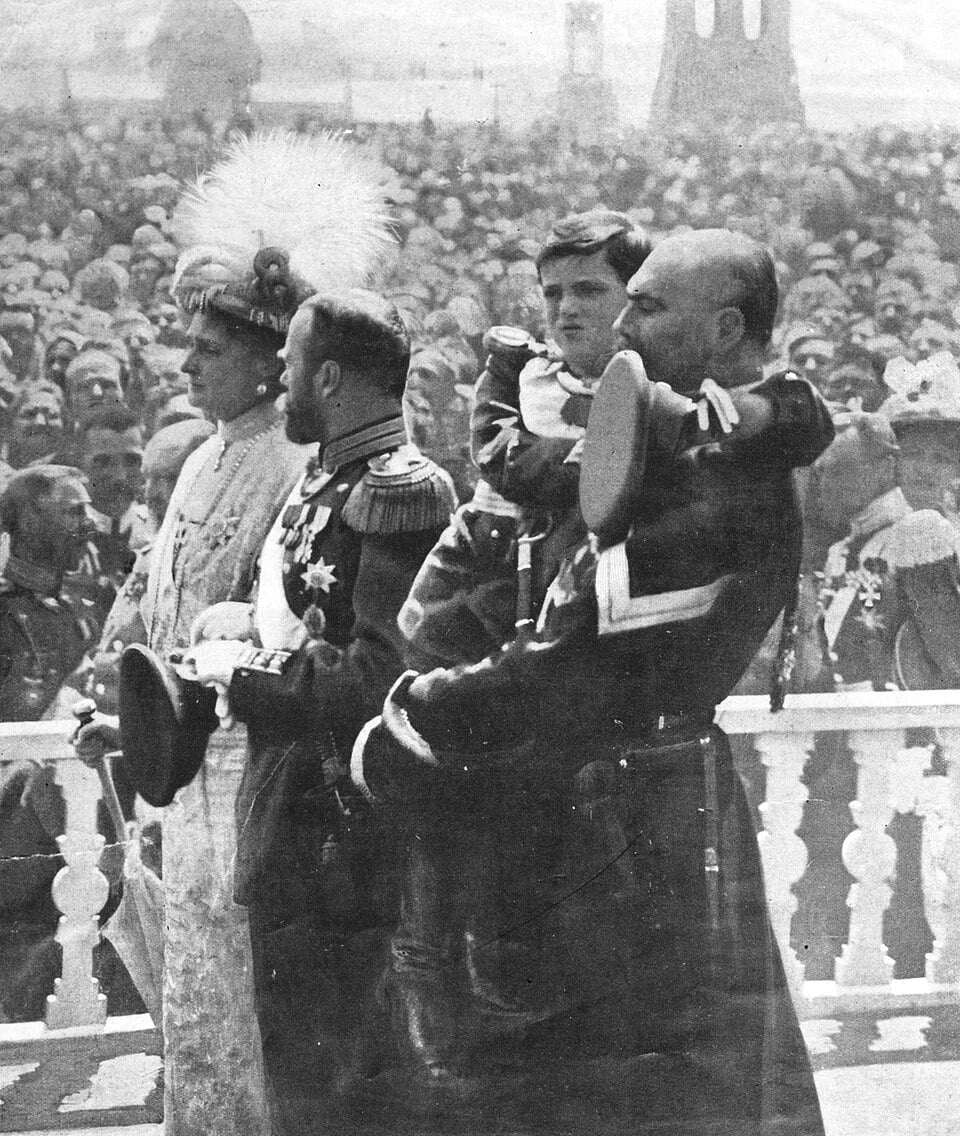Historic black‑and‑white scene of uniformed officers and a hat‑wearing lady on a balcony, facing a dense crowd at a formal public event