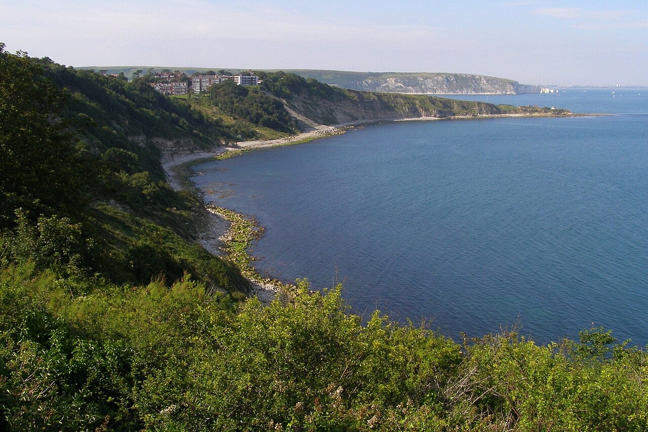 Steep green cliffs curving around a calm blue bay, with rocky shoreline below and a few seaside buildings on the distant headland under a hazy sky