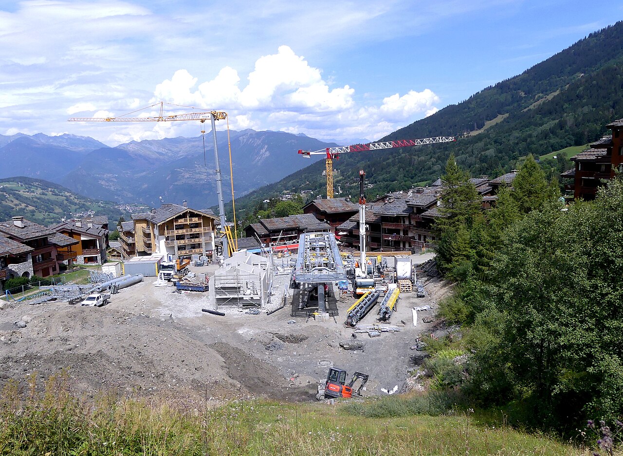 Mountain village construction site with cranes, machinery and new buildings, surrounded by green forested slopes and distant alpine peaks under a blue sky