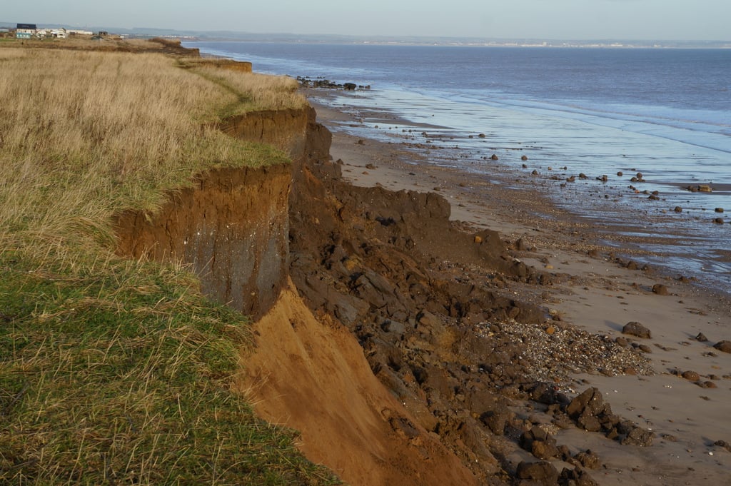 Eroding sandy cliff with grassy top above a wide, rock-strewn beach and calm sea under a clear sky, with distant buildings along the coastline