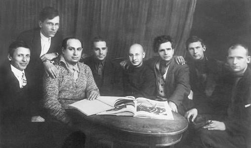 Black-and-white photo of eight men sitting and standing around a small table, looking at an open illustrated book in front of a plain curtain backdrop
