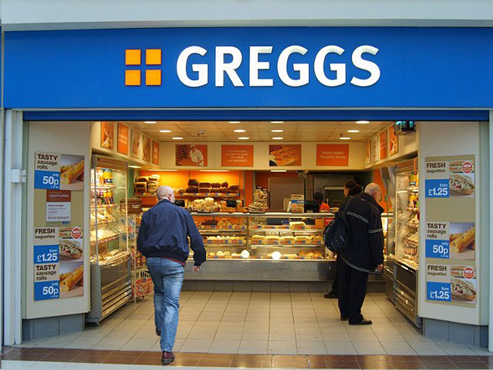 Customers standing at the counter inside a brightly lit Greggs bakery shop, with shelves of pastries and sandwiches and blue price posters at the entrance.