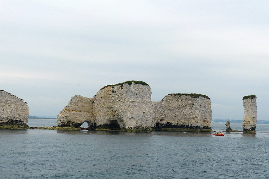 Sea stacks and cliffs of pale rock with grassy tops rise from calm grey water, with a small red boat passing near their base under an overcast sky