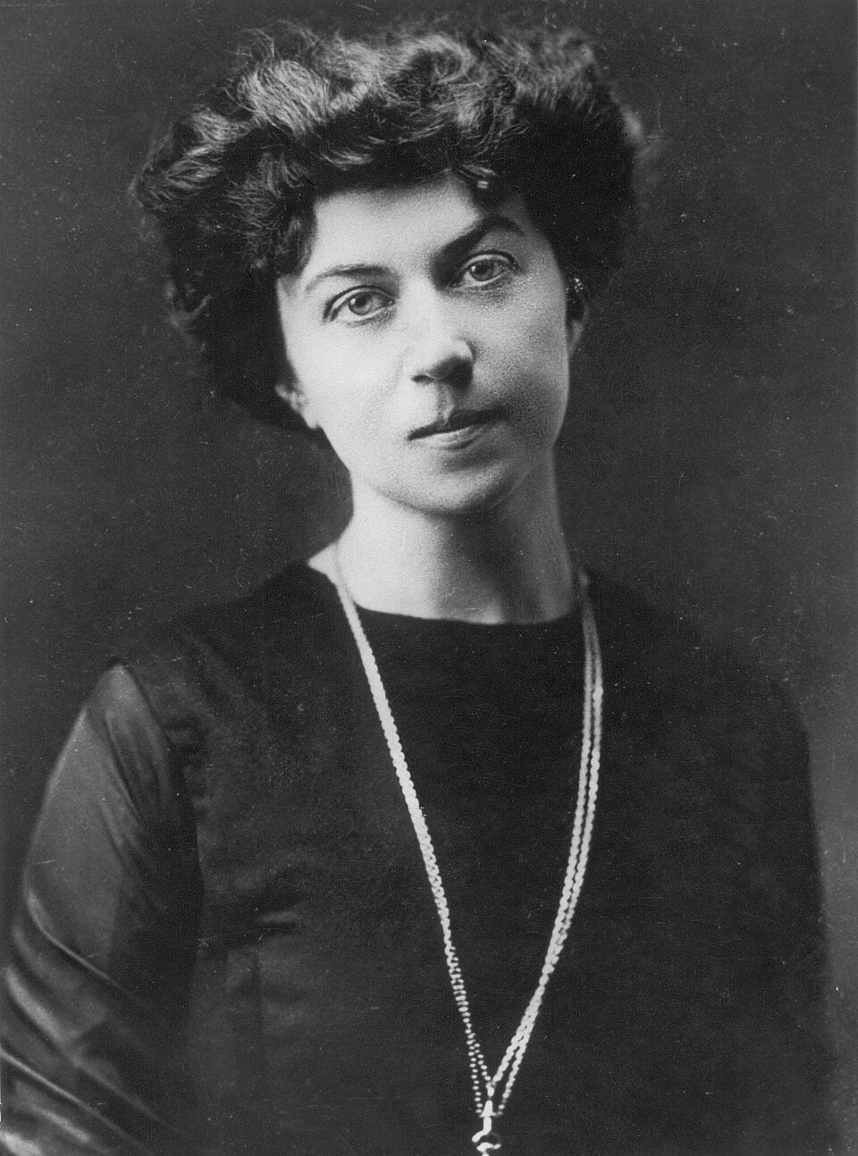 Early 20th-century black-and-white portrait of a young woman with wavy dark hair, wearing a plain dark dress and a long necklace, facing the camera
