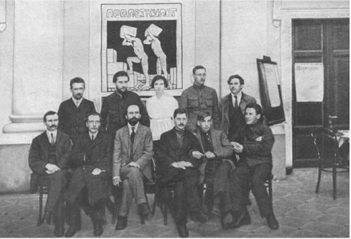 Black-and-white group portrait of early 20th-century men and one woman seated and standing indoors beneath a large Soviet Proletkult workers’ poster