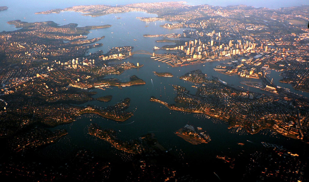 Aerial view of a coastal city at sunset, with skyscrapers, a bridge, islands and peninsulas surrounding a large harbour dotted with boats