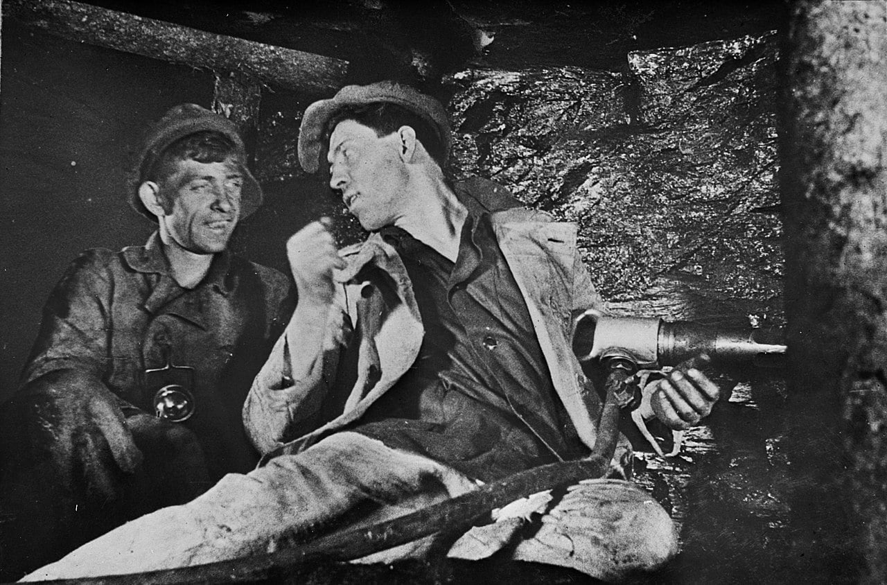 Two coal miners sit in a dark, cramped tunnel, talking as one holds a pneumatic drill against the rough rock wall.