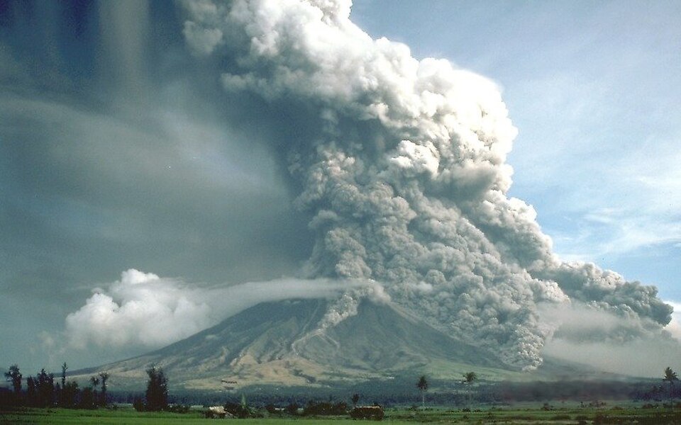 Active volcano erupting with a towering grey ash plume and billowing clouds spreading across the sky above green fields and distant trees