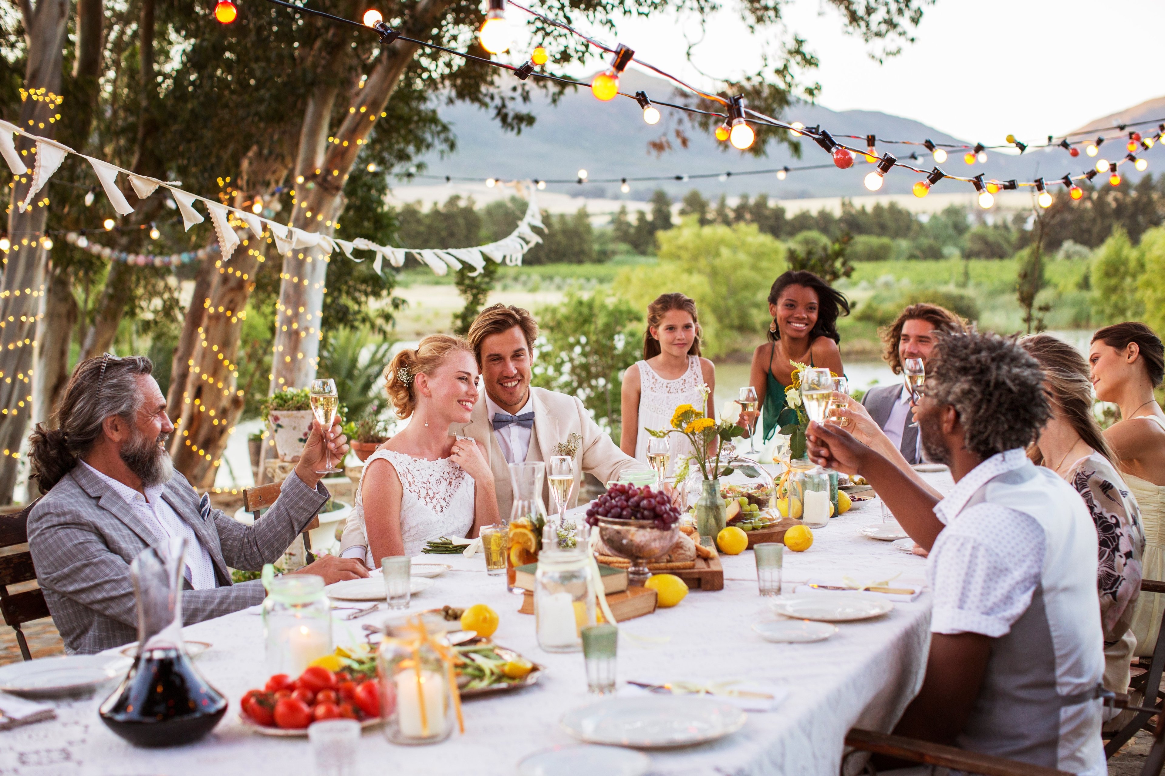 A group of smartly dressed people are at a wedding reception. Some are dancing, others are raising glasses. The atmosphere looks festive and celebratory. A wedding cake is clearly visible.