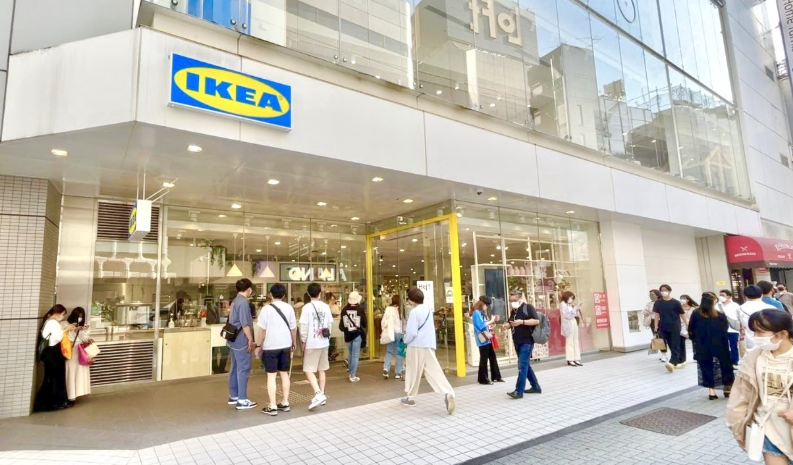 People walking past a busy IKEA shopfront in a modern city street, with glass façade, bright yellow entrance and shoppers entering and leaving