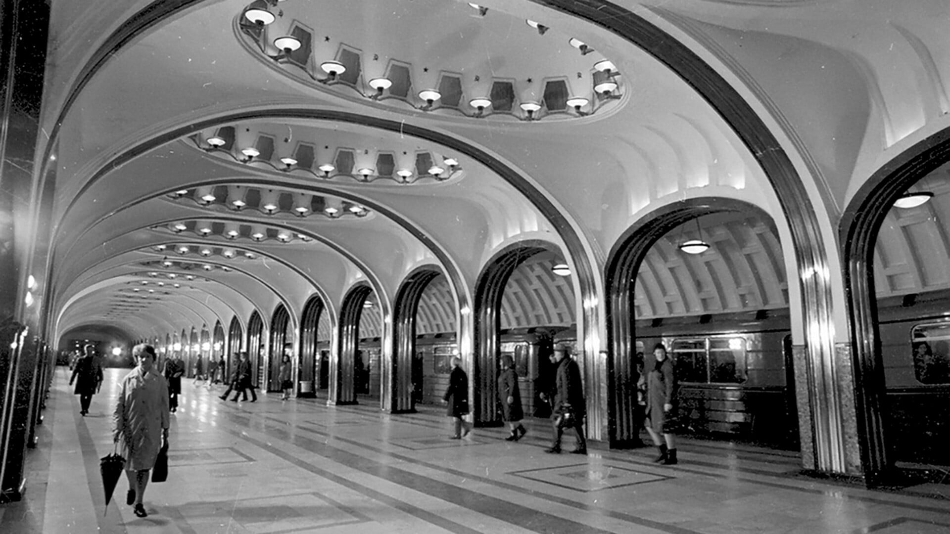 Black-and-white photo of an ornate underground station, with arched ceilings, rows of lights and commuters walking along the polished platform