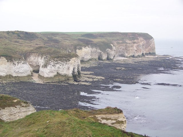 Rugged white sea cliffs with grassy tops overlook a rocky, seaweed-strewn shoreline on a grey, overcast coastal day