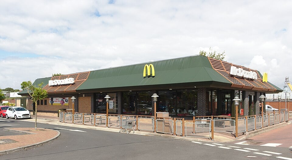 Exterior of a modern McDonald’s restaurant on a street corner, with green roof, large windows, outdoor railings and parked cars nearby