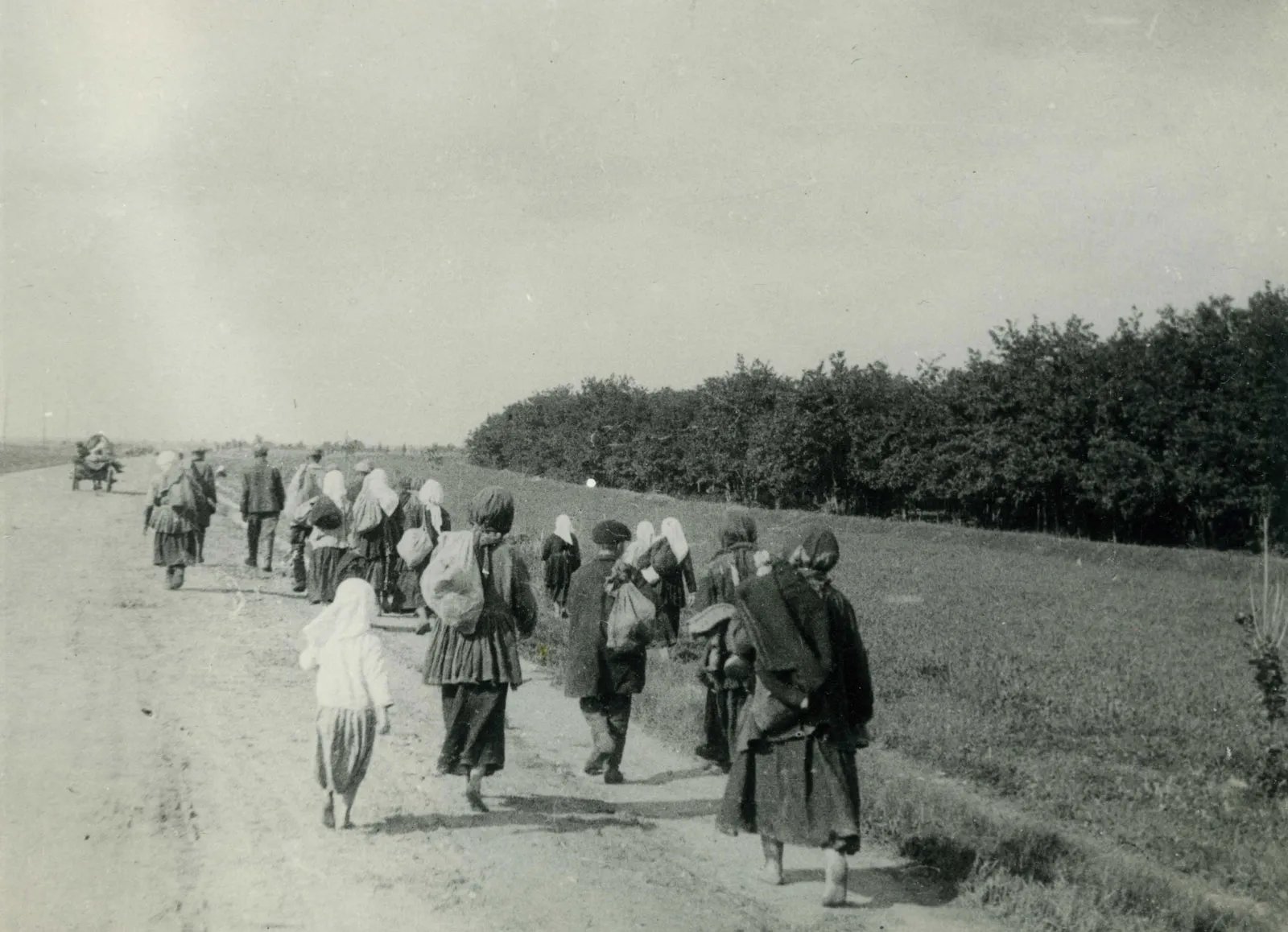 Old black‑and‑white photo of refugees, mostly women and children, walking barefoot along a rural dirt road carrying bundles beside open fields