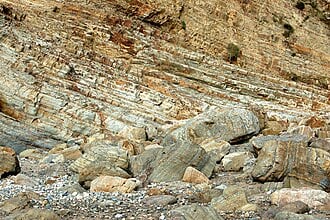Layered, folded rock cliff with visible strata and scattered boulders and pebbles at its base, illustrating coastal sedimentary geology