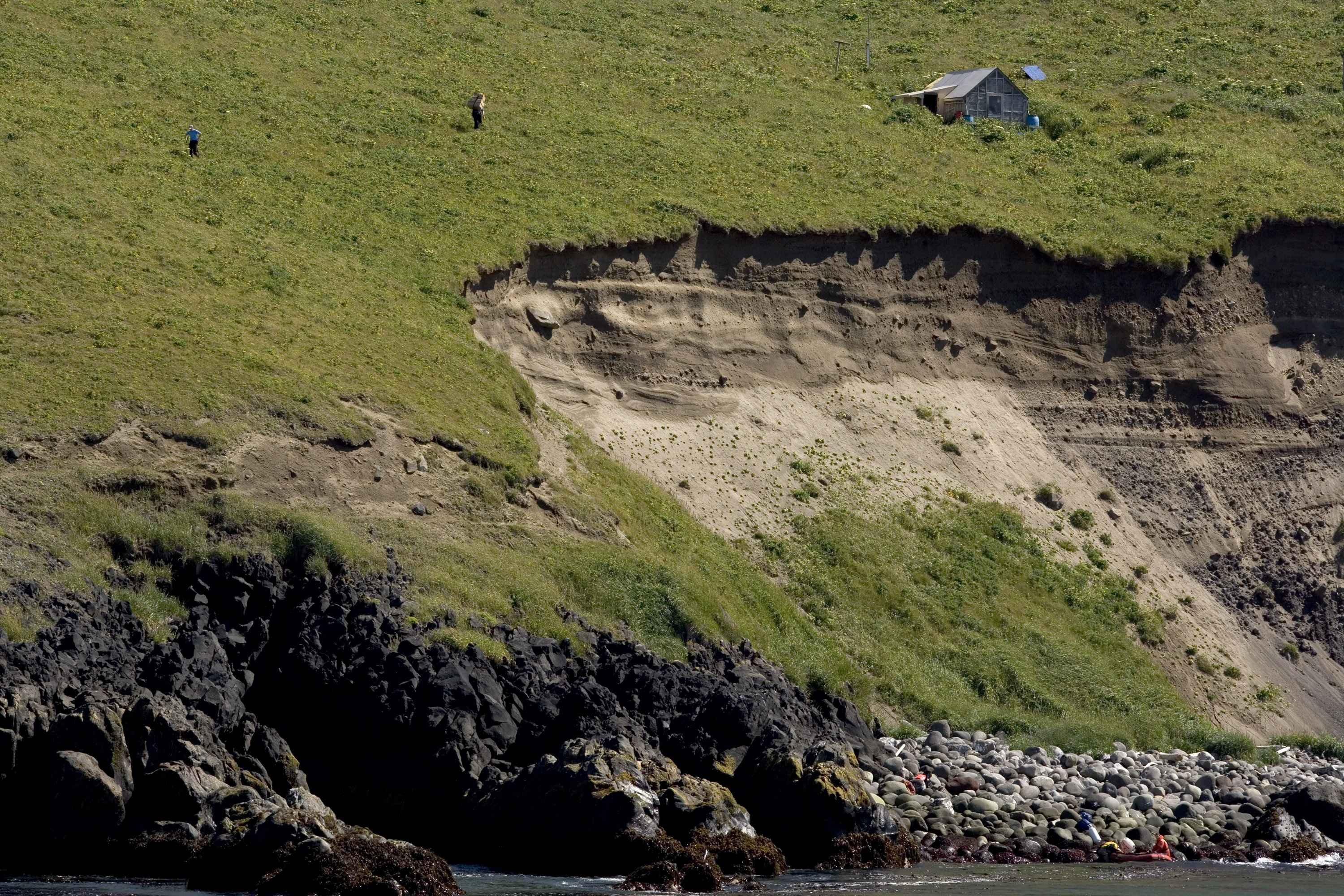 Steep grassy coastal cliff with exposed sandy layers, rocky shoreline below and a small weathered shack on the slope, two people walking across the hillside