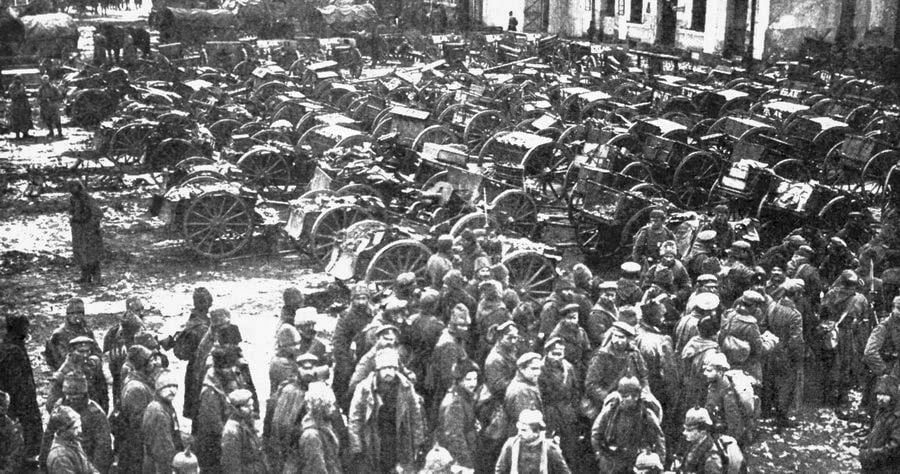 Black‑and‑white wartime scene with rows of abandoned artillery wagons and a large crowd of weary soldiers gathered in a muddy courtyard