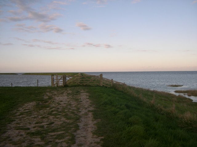 Grassy coastal path leading to a wooden gate on a sea wall, overlooking calm water and a distant low shoreline under a pale evening sky