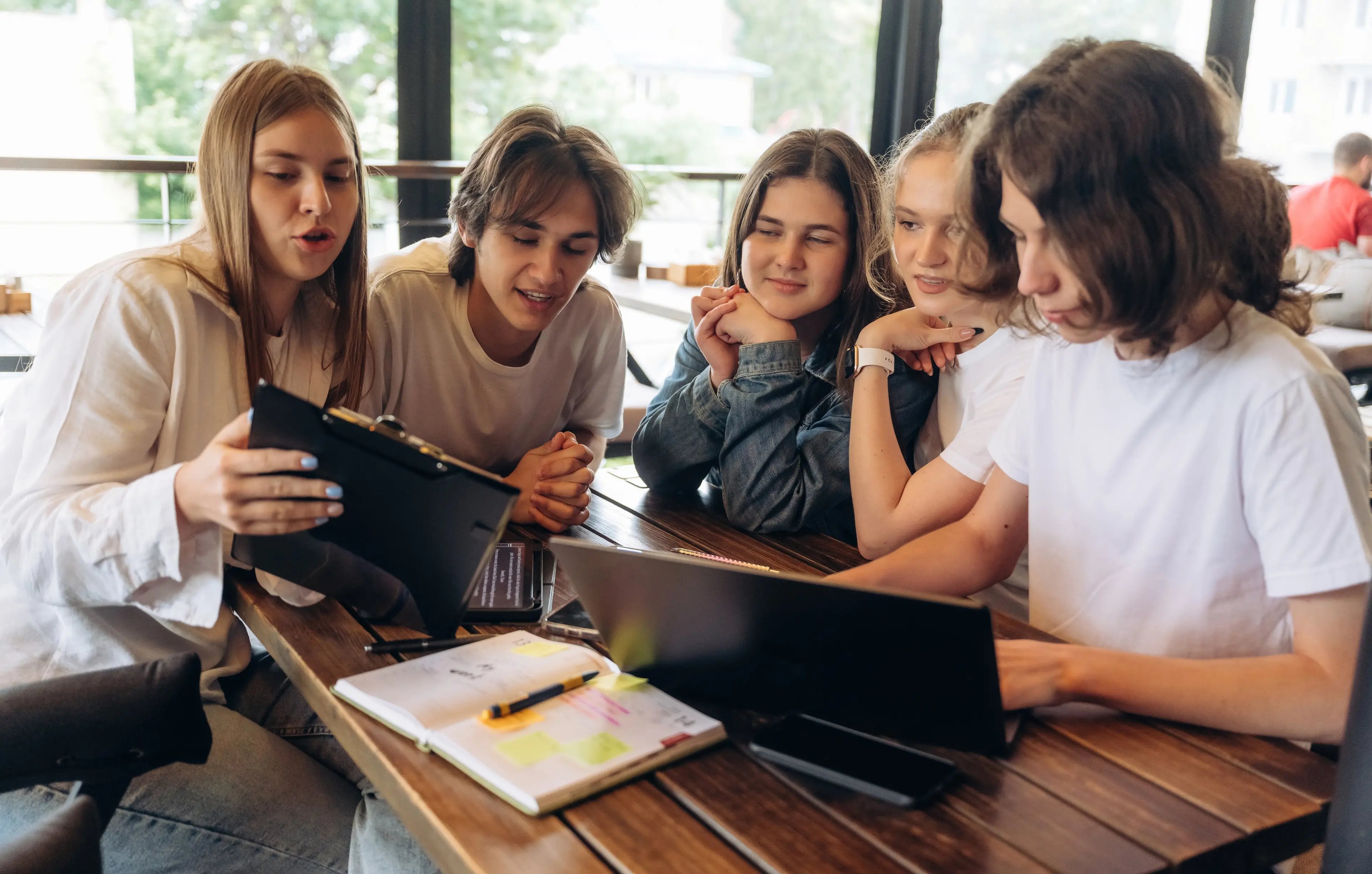 Group of teenagers in casual clothes studying together at a café table with a laptop, tablet and notebook, talking and looking at the screens