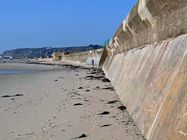 Wide sandy beach beside a tall concrete sea wall on a clear day, with scattered seaweed, distant walkers and low cliffs in the background.