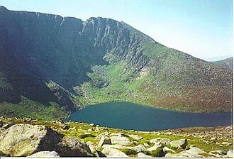 Mountain lake nestled beneath steep rocky cliffs, surrounded by green slopes and scattered foreground boulders under a clear blue sky