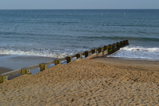 Sandy beach with a wooden groyne stretching into gentle waves under a clear blue sky on a calm day at the seaside