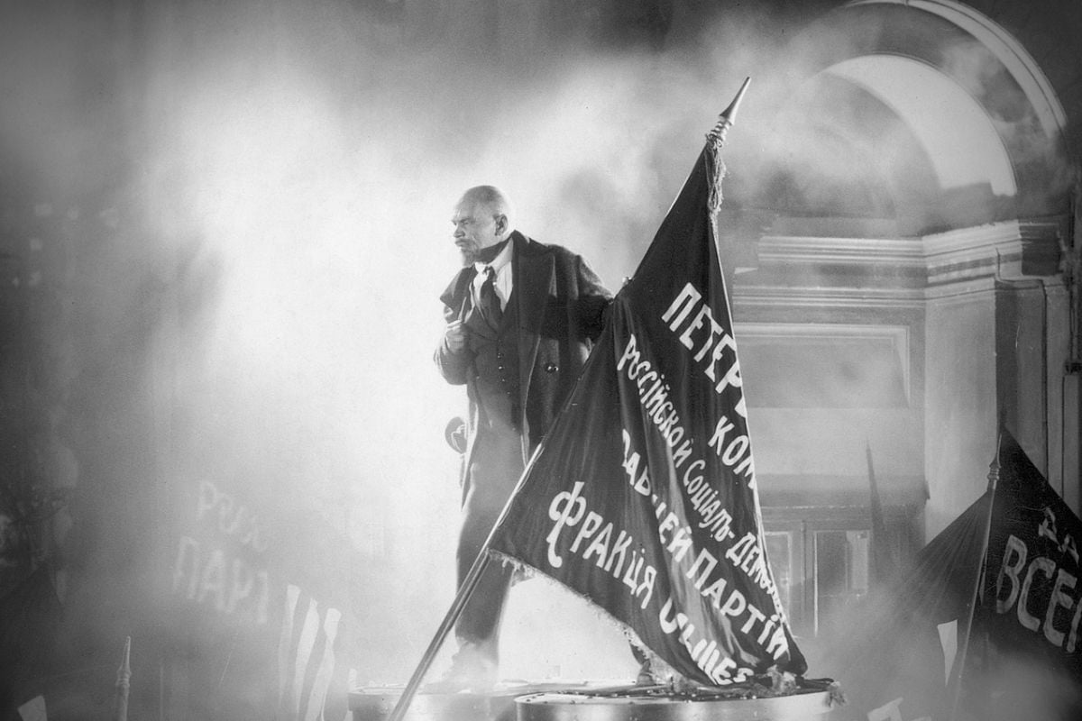 Black-and-white scene of an impassioned orator on a platform, gripping a large Russian banner amid smoke inside a grand, arched building.
