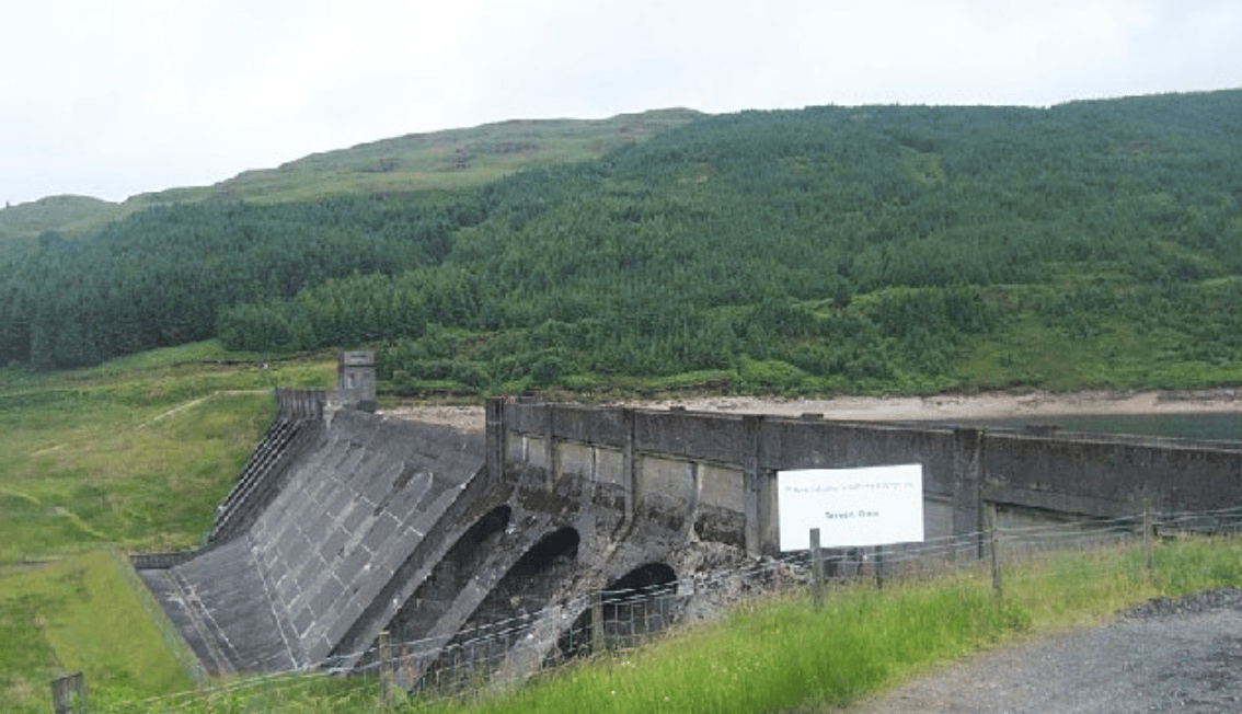 Concrete dam structure in a green, hilly landscape, with dense forest on the slopes and a reservoir partly visible behind the dam wall