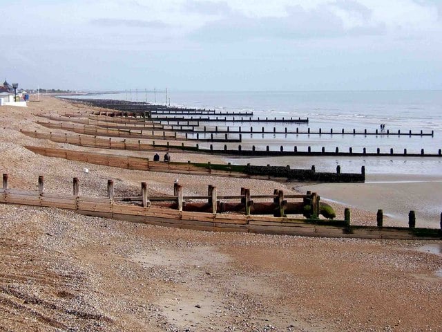 Pebble beach with wooden groynes curving into the calm sea under an overcast sky, a few distant people walking along the shoreline