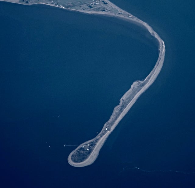 Aerial view of a narrow sandy spit curving into deep blue sea, with a rounded tip and sparse buildings on low land at the distant shoreline