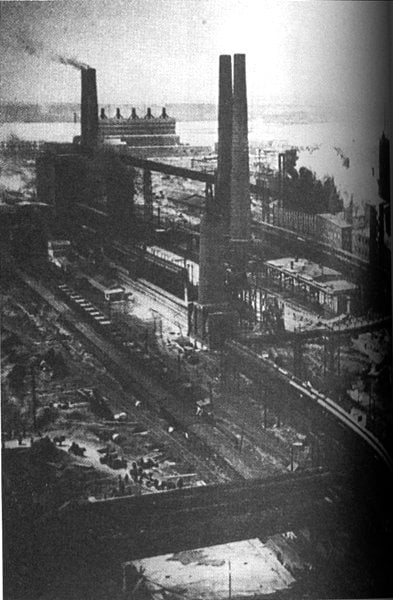 Old black-and-white photograph of a sprawling riverside industrial plant with tall chimneys, smokestacks, rail tracks and heavy machinery in a smoky atmosphere